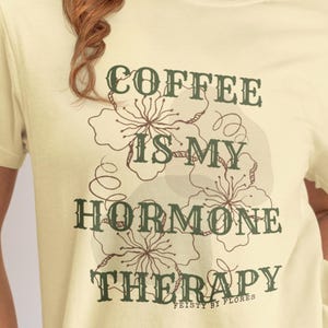 May include: A light yellow t-shirt with the text "COFFEE IS MY HORMONE THERAPY" in dark green. The shirt features a floral design and is from the brand "FEISTY BY FLORES".