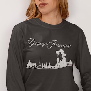 May include: Dark gray long-sleeve shirt with the text "Divine Feminine" in a script font. A white graphic of a woman in a hat and a city skyline is below the text. The shirt is made of a soft material.
