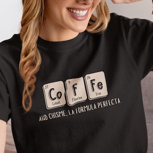 Peut inclure: T-shirt noir avec un motif de tableau p&eacute;riodique &eacute;pelant "COFFEE" avec les &eacute;l&eacute;ments Cobalt, Fluorine et Fer. Le texte "AND CHISME, LA FORMULA PERFECTA" est en dessous. Une casquette de baseball beige est port&eacute;e.