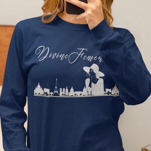 May include: Navy blue long-sleeve shirt with the text "Divine Femen" in a cursive font. A white graphic of a cityscape and a woman in a hat is below the text. The shirt is made of a soft, comfortable material.