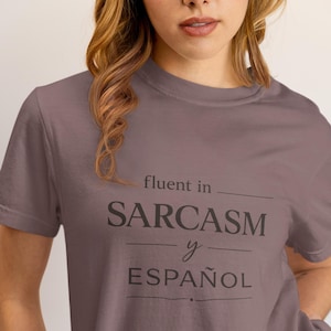 Może przedstawiać: Mauve koszulka z czarnym napisem "fluent in SARCASM y ESPA&Ntilde;OL". Koszulka z kr&oacute;tkim rękawem wydaje się być wykonana z miękkiego materiału. Modelka ma na sobie jasnor&oacute;żowe spodnie.