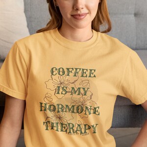 May include: Mustard yellow t-shirt with the text "COFFEE IS MY HORMONE THERAPY" in a green, vintage-style font. The shirt features a floral design. The text "PAISTI BY FLORES" is below the main text.