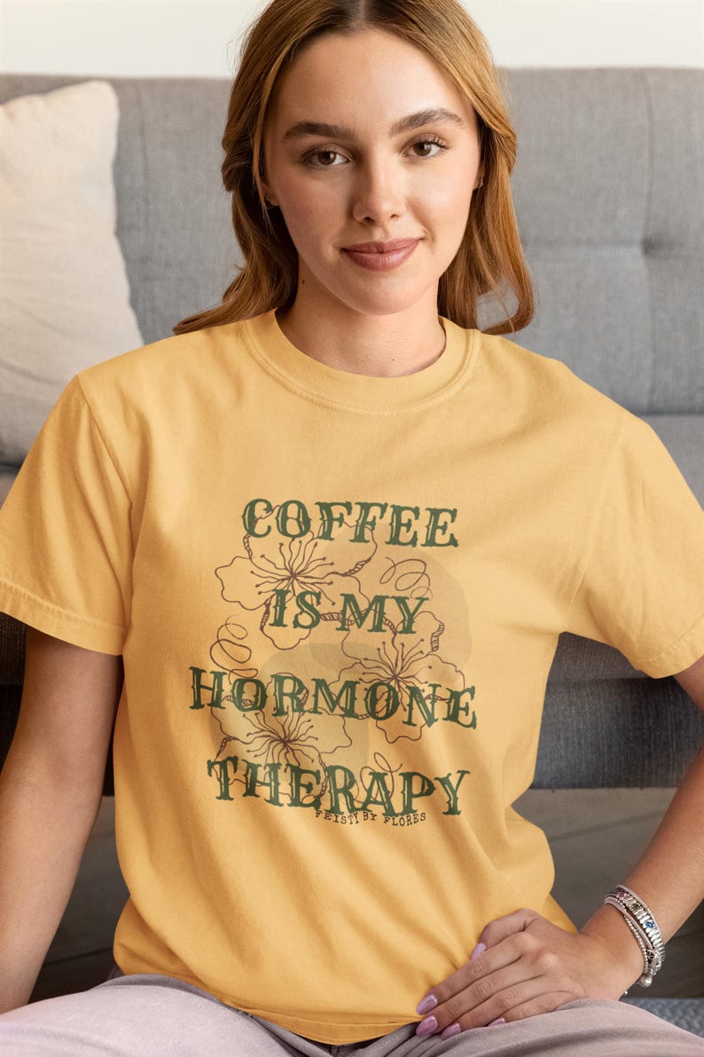 May include: Mustard yellow t-shirt with the text "COFFEE IS MY HORMONE THERAPY" in a green, vintage-style font. The shirt features a floral design. The text "PAISTI BY FLORES" is below the main text.