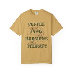 May include: Mustard yellow t-shirt with the text "COFFEE IS MY HORMONE THERAPY" in dark green, along with floral illustrations. The shirt is a casual wear item, perfect for everyday use.