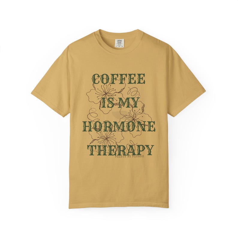 May include: Mustard yellow t-shirt with the text "COFFEE IS MY HORMONE THERAPY" in dark green, along with floral illustrations. The shirt is a casual wear item, perfect for everyday use.