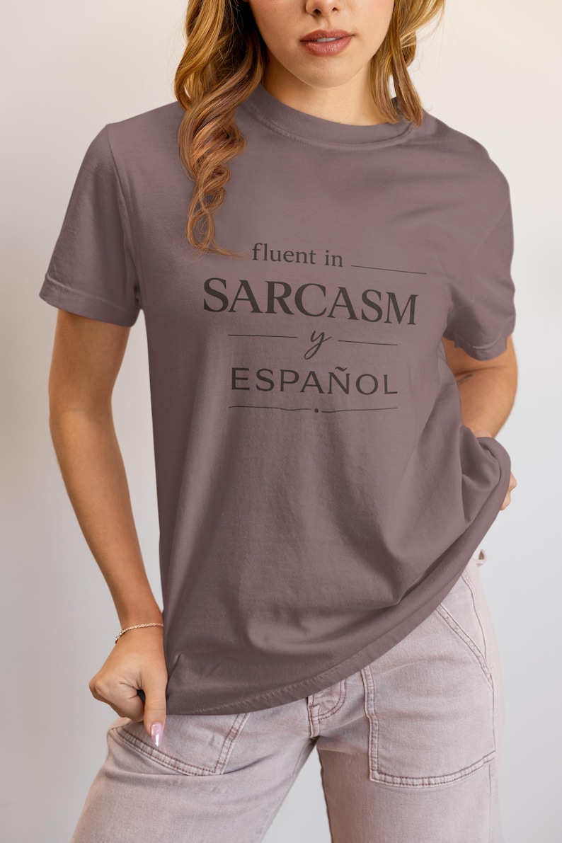 Może przedstawiać: Mauve koszulka z czarnym napisem "fluent in SARCASM y ESPA&Ntilde;OL". Koszulka z kr&oacute;tkim rękawem wydaje się być wykonana z miękkiego materiału. Modelka ma na sobie jasnor&oacute;żowe spodnie.