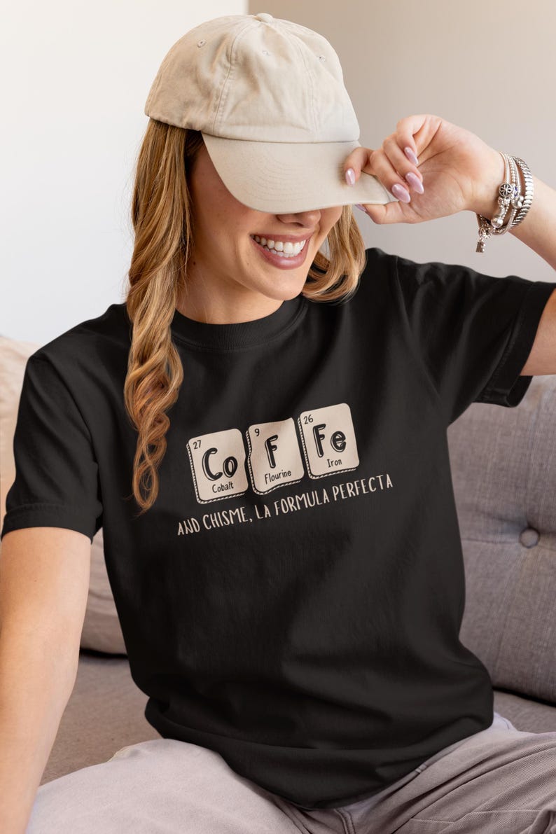Peut inclure: T-shirt noir avec un motif de tableau p&eacute;riodique &eacute;pelant "COFFEE" avec les &eacute;l&eacute;ments Cobalt, Fluorine et Fer. Le texte "AND CHISME, LA FORMULA PERFECTA" est en dessous. Une casquette de baseball beige est port&eacute;e.