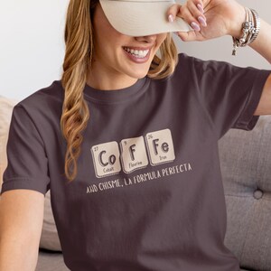Peut inclure: T-shirt violet fonc&eacute; avec le mot "COFFEE" &eacute;pel&eacute; avec des &eacute;l&eacute;ments du tableau p&eacute;riodique. Le t-shirt porte &eacute;galement l'inscription "AND CHISME, LA FORMULA PERFECTA" en dessous.