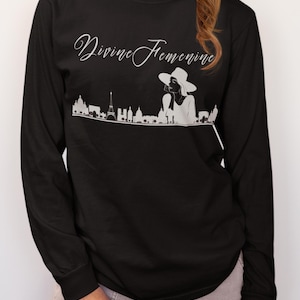 May include: Black long-sleeve shirt with the text "Divine Feminine" in cursive script. A white graphic of a woman in a hat and a city skyline is below the text. The shirt is made of a soft material.
