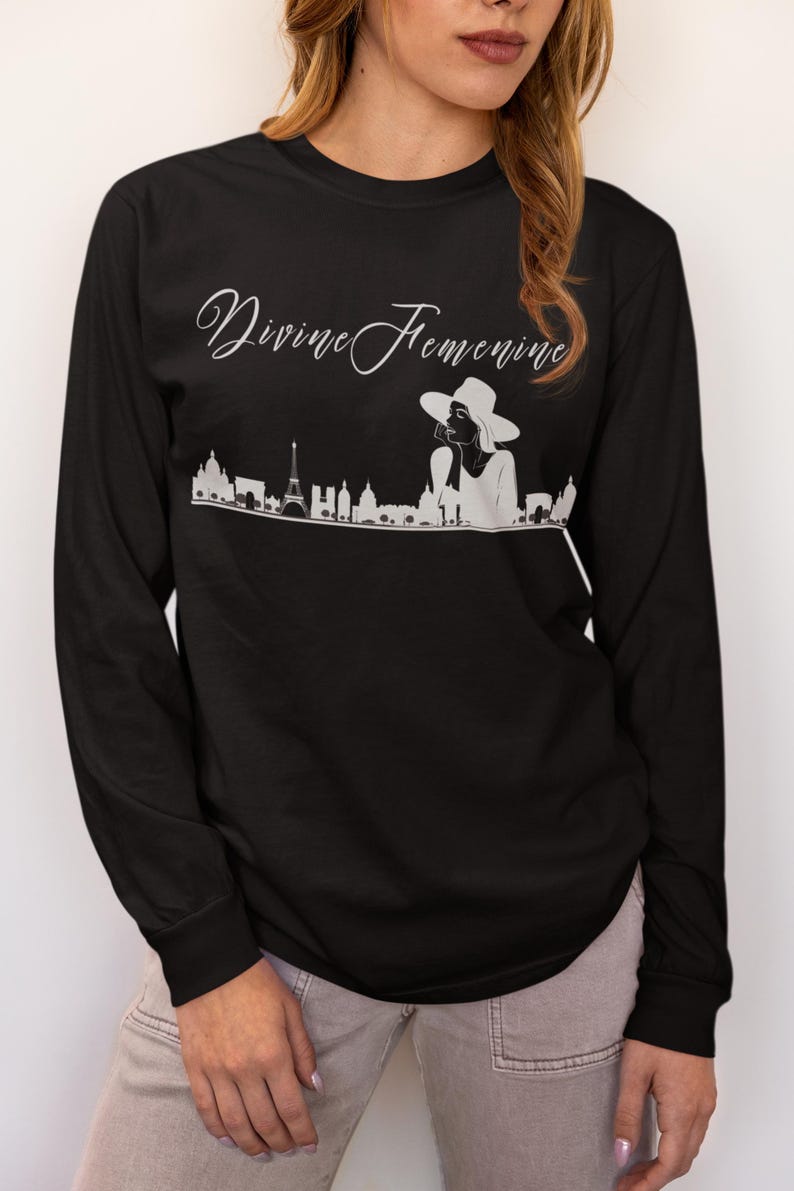 May include: Black long-sleeve shirt with the text "Divine Feminine" in cursive script. A white graphic of a woman in a hat and a city skyline is below the text. The shirt is made of a soft material.