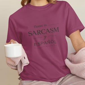 Może przedstawiać: Bordowa koszulka z napisem "fluent in SARCASM y ESPA&Ntilde;OL" w ciemnoszarym kolorze. Koszulka jest trzymana przez osobę, kt&oacute;ra trzyma r&oacute;wnież biały kubek. Osoba ta ma na sobie r&oacute;żowy sweter.