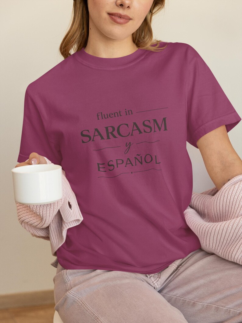 Może przedstawiać: Bordowa koszulka z napisem "fluent in SARCASM y ESPA&Ntilde;OL" w ciemnoszarym kolorze. Koszulka jest trzymana przez osobę, kt&oacute;ra trzyma r&oacute;wnież biały kubek. Osoba ta ma na sobie r&oacute;żowy sweter.