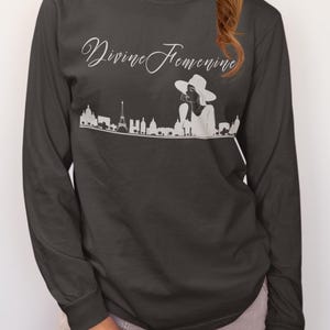 May include: Dark gray long-sleeve shirt with the text "Divine Feminine" in a cursive font. Below the text is a white graphic of a cityscape with a woman wearing a hat. The shirt is a crew neck.