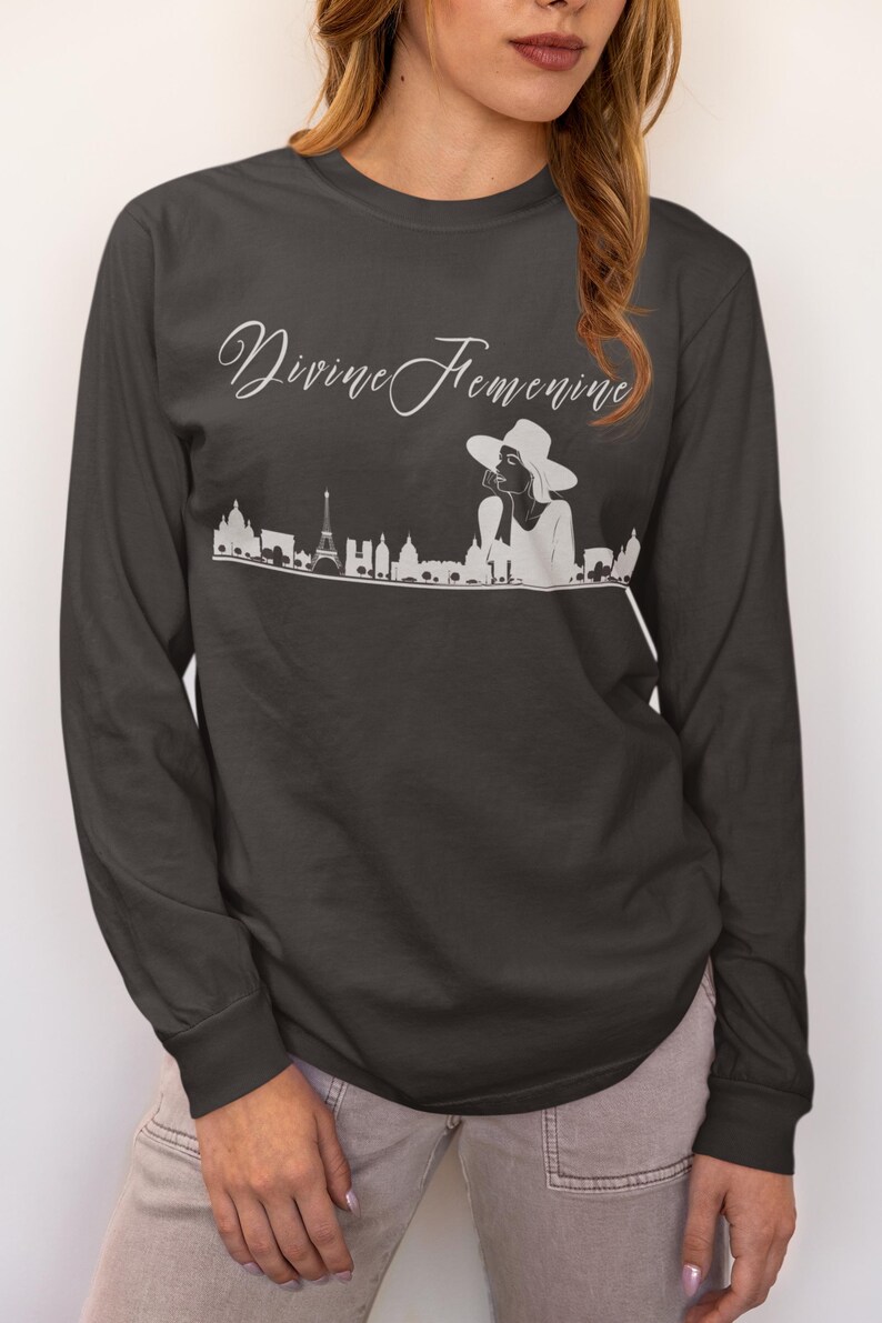 May include: Dark gray long-sleeve shirt with the text "Divine Feminine" in a cursive font. Below the text is a white graphic of a cityscape with a woman wearing a hat. The shirt is a crew neck.