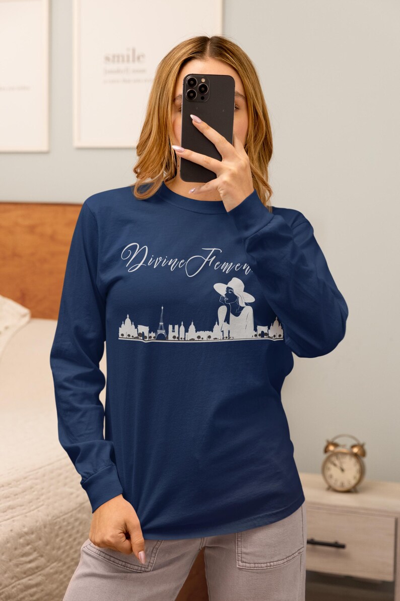 May include: Navy blue long-sleeve shirt with the text "Divine Femen" in a cursive font. A white graphic of a cityscape and a woman in a hat is below the text. The shirt is made of a soft, comfortable material.