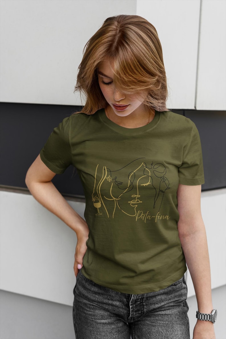 May include: Olive green t-shirt featuring a gold line art design of a woman's face, a wine glass, and the text "Spita-fina". The design is centered on the front of the shirt. The model is wearing dark wash jeans.