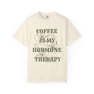 May include: Cream-colored t-shirt with the text "COFFEE IS MY HORMONE THERAPY" in dark green, along with floral illustrations. The shirt is a casual, short-sleeved garment, perfect for everyday wear.