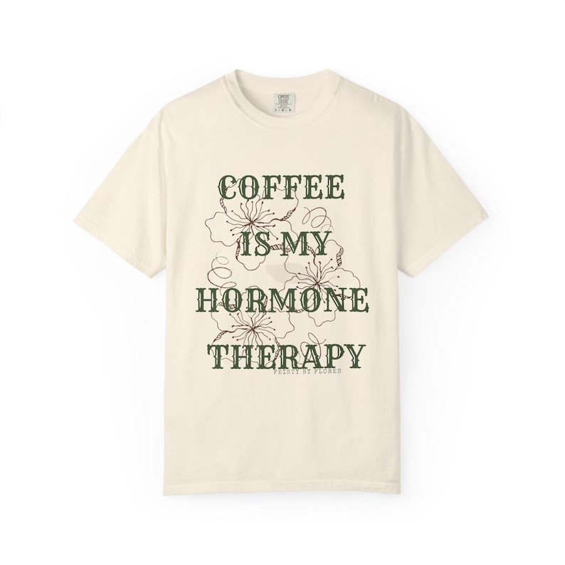 May include: Cream-colored t-shirt with the text "COFFEE IS MY HORMONE THERAPY" in dark green, along with floral illustrations. The shirt is a casual, short-sleeved garment, perfect for everyday wear.