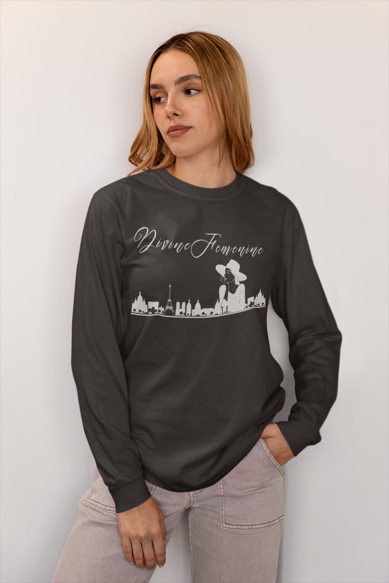 May include: Dark gray long-sleeve shirt with the text "Divine Feminine" in a script font. A white graphic of a woman in a hat and a city skyline is below the text. The shirt is made of a soft material.