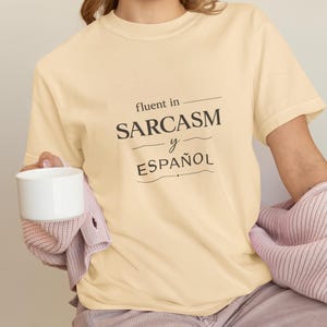 Może przedstawiać: Jasnoż&oacute;łta koszulka z czarnym napisem "fluent in SARCASM y ESPA&Ntilde;OL". Osoba trzyma biały kubek i ma na sobie r&oacute;żowy sweter.