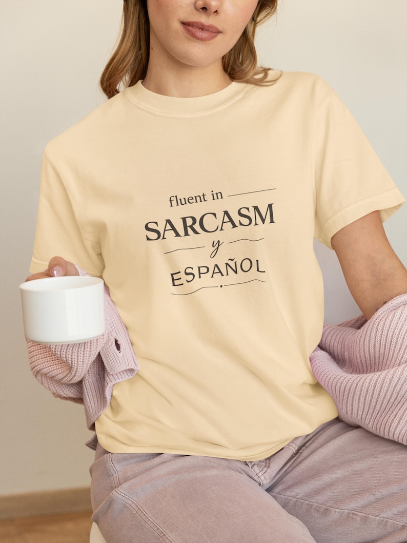 Może przedstawiać: Jasnoż&oacute;łta koszulka z czarnym napisem "fluent in SARCASM y ESPA&Ntilde;OL". Osoba trzyma biały kubek i ma na sobie r&oacute;żowy sweter.