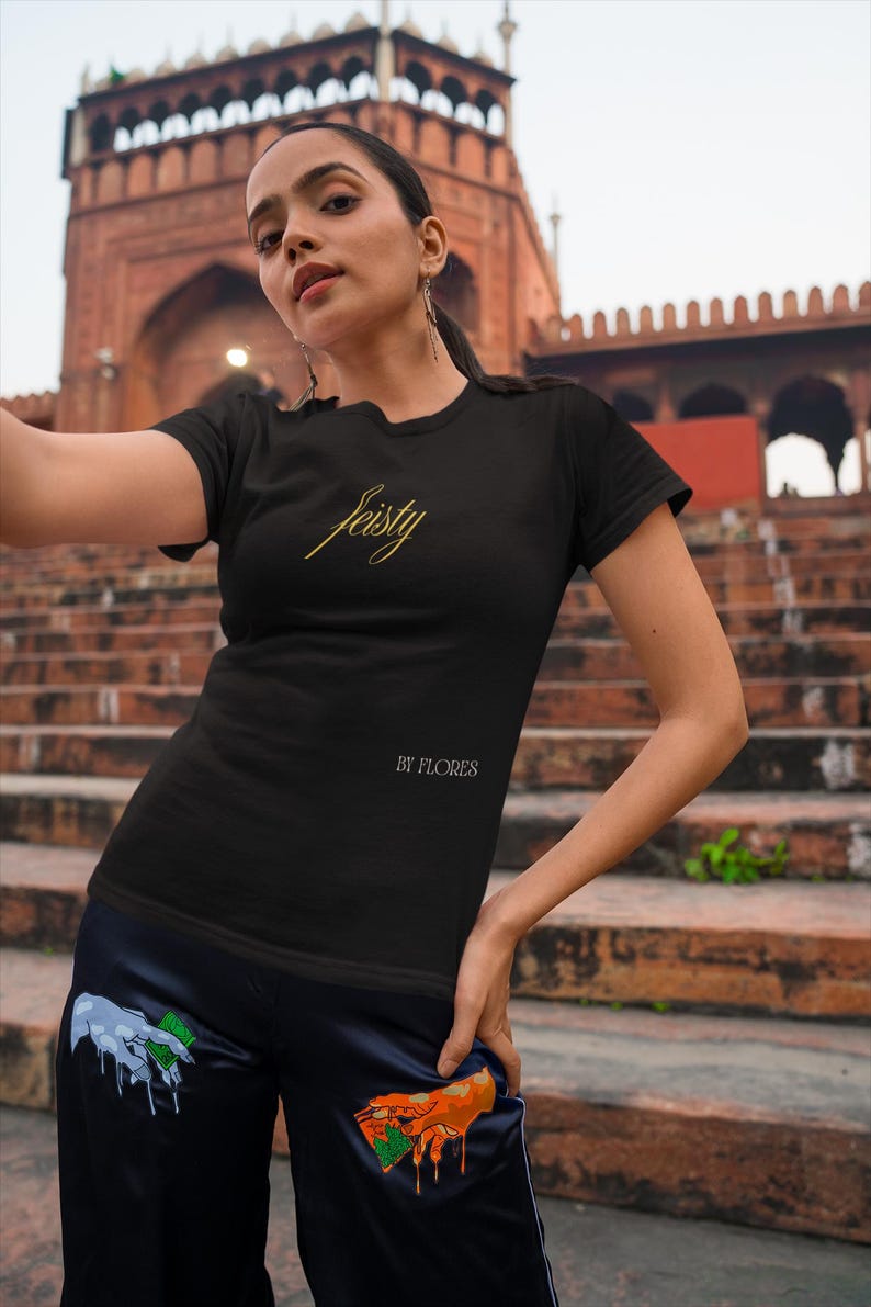 May include: Black t-shirt with the word "feisty" in gold script. The shirt also has the text "BY FLORES" below the word "feisty". The model is wearing black pants with colorful hand graphics.