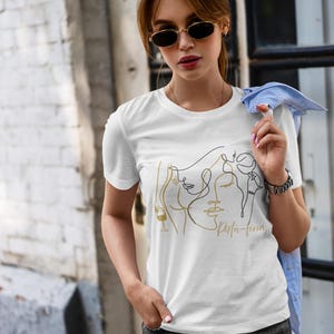 May include: White t-shirt featuring a gold and black line art design of faces and a wine glass. The text "Spirita-fina" is printed in gold. The shirt has short sleeves.