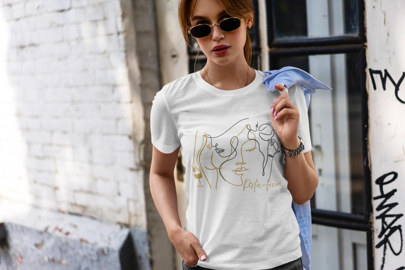May include: White t-shirt featuring a gold and black line art design of faces and a wine glass. The text "Spirita-fina" is printed in gold. The shirt has short sleeves.