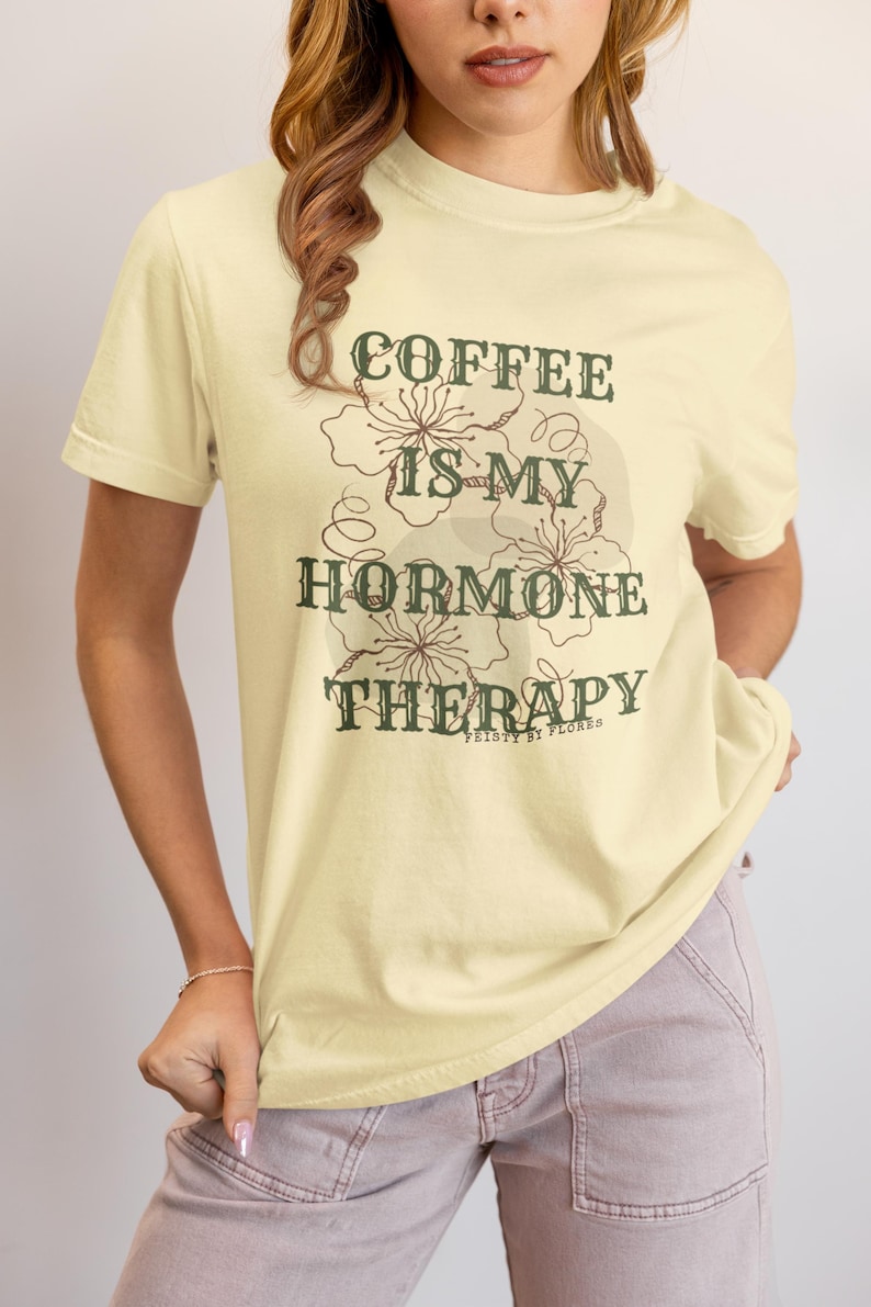 May include: A light yellow t-shirt with the text "COFFEE IS MY HORMONE THERAPY" in dark green. The shirt features a floral design and is from the brand "FEISTY BY FLORES".