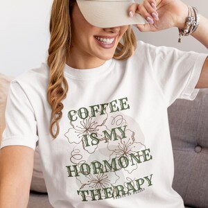 May include: White t-shirt with the text "COFFEE IS MY HORMONE THERAPY" in green and brown. The shirt features floral designs and is worn by a person wearing a beige baseball cap.