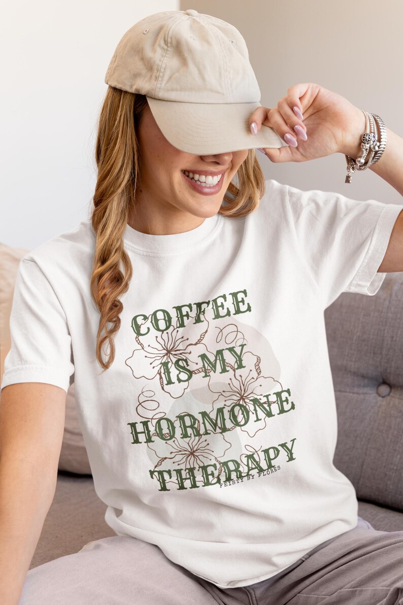 May include: White t-shirt with the text "COFFEE IS MY HORMONE THERAPY" in green and brown. The shirt features floral designs and is worn by a person wearing a beige baseball cap.