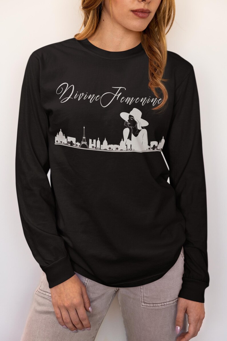 May include: Black long-sleeve shirt with the text "Divine Feminine" in cursive script. A white graphic of a woman in a hat and a city skyline is below the text. The shirt is made of a soft material.