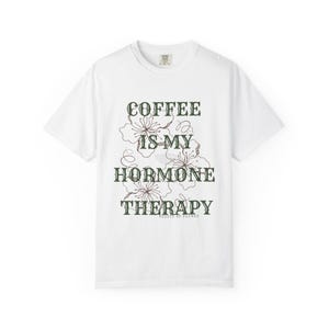 May include: White crew neck t-shirt with the text "COFFEE IS MY HORMONE THERAPY" in dark green, with floral accents. The shirt is a casual style, perfect for everyday wear. The text is in a decorative font.