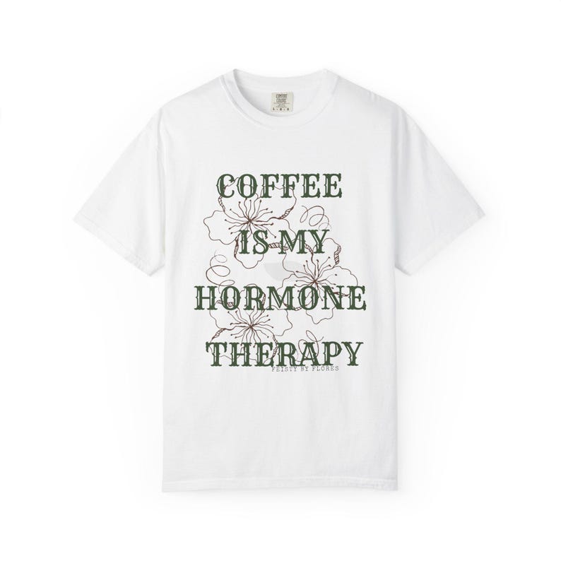 May include: White crew neck t-shirt with the text "COFFEE IS MY HORMONE THERAPY" in dark green, with floral accents. The shirt is a casual style, perfect for everyday wear. The text is in a decorative font.