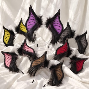 May include: A collection of headbands featuring pointed, bat-like ears covered in black faux fur. The inner ear sections are made of colored fabric in shades of purple, yellow, pink, red, grey, and brown, each with a ribbed, textured pattern.