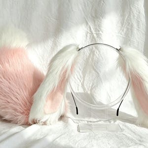 May include: A set of faux fur animal ears on a thin headband and a matching fluffy tail. The ears and tail feature long, soft white fur on the outer edges with light pink fur accents on the inner sections and a pink base on the tail.