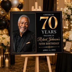 May include: A rectangular black birthday sign on a wooden easel featuring a large gold 70, the text Cheers to 70 Years, Welcome to Robert Johnson's 70th Birthday, April 14, 20XX, and a portrait of a man in a black suit jacket and shirt.