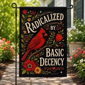 May include: A black garden flag featuring a red cardinal bird perched on a branch, surrounded by red and yellow flowers and green foliage. The text reads RADICALIZED BY BASIC DECENCY in a decorative cream-colored font with small star accents.
