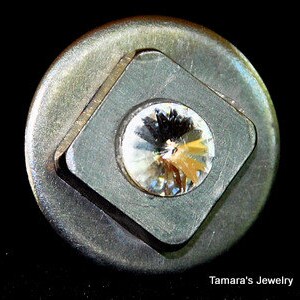 Könnte beinhalten: Ein silberfarbener Knopf mit einem quadratischen Rahmen und einem klaren Kristall in der Mitte.