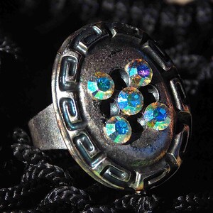 Könnte beinhalten: Ein silberner Ring mit einem großen, runden, schwarzen Einsatz, der mit schillernden blauen Strasssteinen besetzt ist. Der Einsatz hat ein dekoratives, geometrisches Muster.