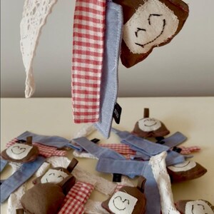 May include: A collection of small fabric ornaments featuring brown plush shapes with embroidered smiling faces, attached to various fabric strips including red and white gingham, solid light blue, and white lace, all suspended from a pink loop.