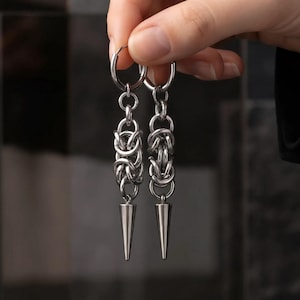 May include: A pair of silver-toned drop earrings featuring a circular hoop top, a central section of intricate interlocking chainmail links, and a smooth, pointed conical spike at the bottom. The earrings are crafted from polished metal.