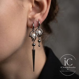 Könnte beinhalten: Ein Paar silberfarbene Ohrringe mit einem Metallkäfig-Design, das eine weiße Perle umschließt. Ein Ohrring hat einen langen, spitzen Anhänger, während der andere eine kleinere hängende Kugel aufweist. Beide Ohrringe hängen an einem Silberring.