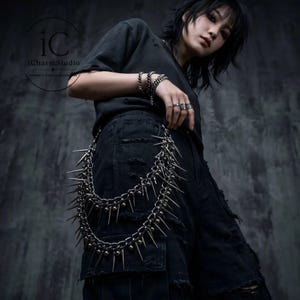 May include: A silver-colored, spiked chain belt with multiple rows of sharp spikes and small ball accents. The belt is worn over black shorts. The image also shows a black t-shirt and multiple silver bracelets and rings. The iCharmStudio logo is visible.
