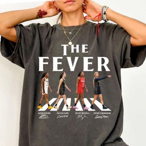 May include: A charcoal gray t-shirt featuring the text THE FEVER in white capital letters above a graphic of four basketball players walking across a crosswalk. Each player is identified by name below their image: Aliyah Boston, Caitlin Clark.