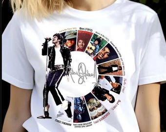 May include: White t-shirt featuring a graphic of Michael Jackson performing on stage, surrounded by album covers and a signature. The design includes album titles and dates, creating a circular collage.