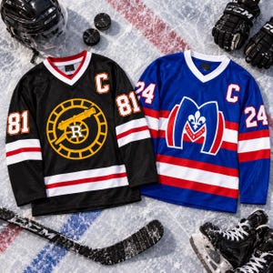 May include: Two hockey jerseys, one black with a yellow cannon emblem and number 81, the other blue with a red and white M logo and number 24. Both feature a white V-neck collar, captain C patch, and striped sleeves, surrounded by hockey gear.