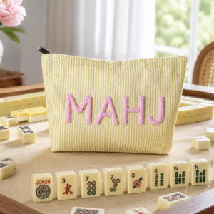 Può includere: Una pochette in tessuto a righe gialle e bianche con le lettere MAHJ ricamate in rosa sul davanti. La pochette ha una chiusura lampo argentata ed è posizionata tra diverse tessere del mahjong color crema su una superficie.