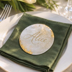 May include: A circular white mother-of-pearl place card featuring the name Alice written in gold script. The edges of the disc are accented with textured gold leaf detailing, creating a reflective and metallic finish on the natural shell surface.
