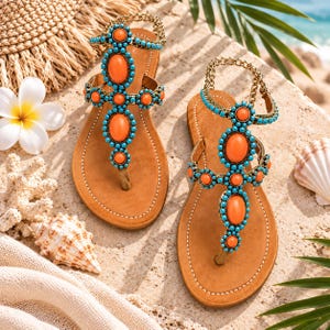 May include: A pair of tan leather thong sandals featuring a vertical arrangement of large, smooth orange oval stones surrounded by small, bright turquoise beads. The sandals include a gold-tone chain ankle strap and white stitching along the sole.